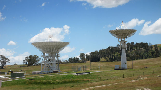 Two antennas in a field with blue sky. Left antenna Deep Space Station 34, right antenna the smaller Deep Space Sation 33