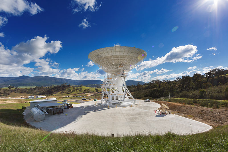 Wide shot showing DSS35 in the centre facing up, very blue sky, concrete hardstand around the antenna, buildings to the left and green grass