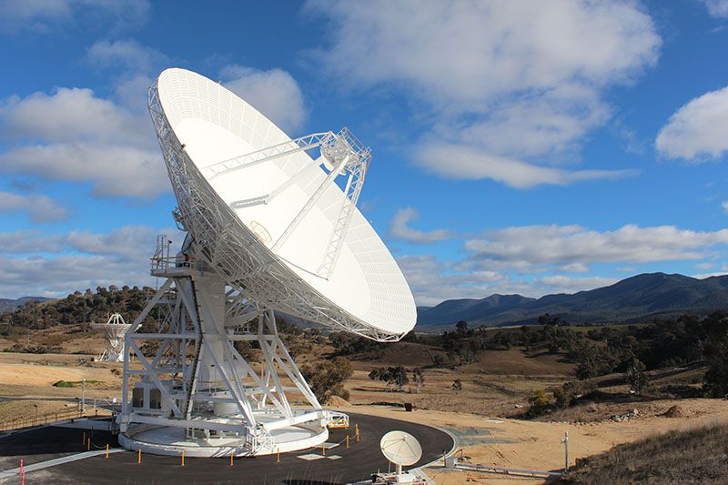 DSS36 on left, on an angle facing to the right. Blue sky with clouds, hills in the background, tiny dish to the right