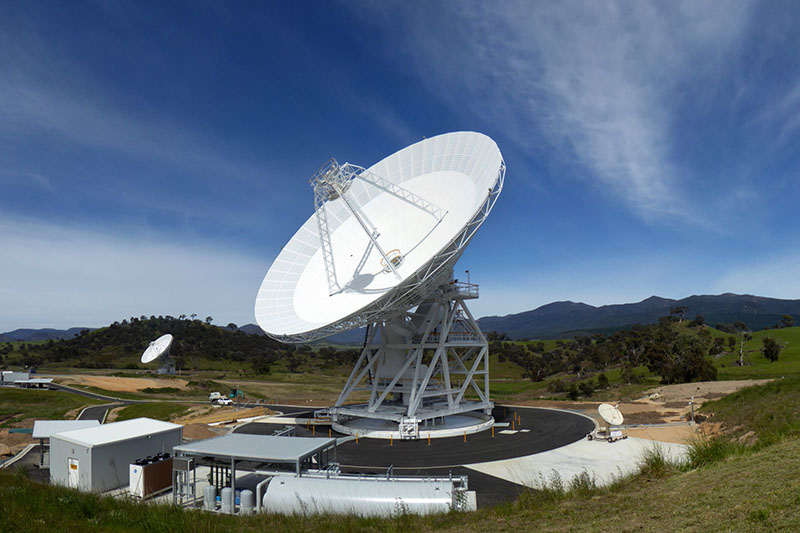 Wider shot showing DSS36 in the centre, on an angle facing to the left. DSS45 in the background to the left and tiny dish to the right. Blue sky with clouds and hills in the background