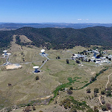 Side on aerieal view of the Canberra Deep Space Communication Complex, showing six antennas, buildings, hills and the blue sky in the background.
