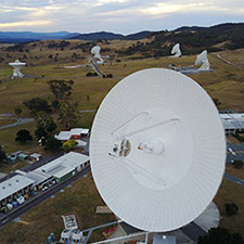 Low aerial view of the Canberra Deep Space Communication Complex, with DSS43 large in the foreground on an angle, and four antennas in the background, along with hills and cloudy sky.