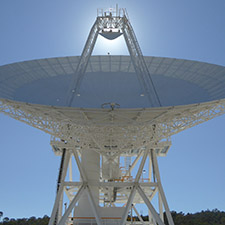 Most of the structure of Deep Space Station 34, dishing facing almost straight up with the Sun behind the subreflector at the top of the quad legs. Blue sky.