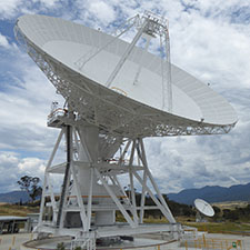 Deep Space Station 35 on an angle showing the quad legs and subreflector with hills, grass, another antenna in the background and cloudy sky.
