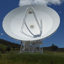 Deep Space Station 35 showing the dish, quad legs and subreflector, green grass in front, hills behind and a blue sky with clouds.