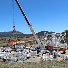 Sections of the pedestal structure of Deep Space Station 35 on the ground with a crane attached to part of it, hills and blue sky in the background.