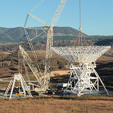 Crane attached to the dish of Deep Space Station 35 with the dish now sitting on the pedestal. Blue sky and hills behind, a yellow glow on the ground from the rising Sun.