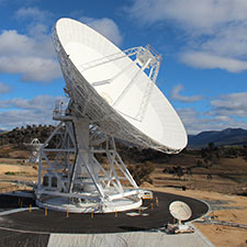 Deep Space Station 36 on an angle lit up by the Sun, bright blue sky with clouds and hills behind.