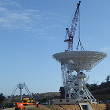 Crane lowering the dish of Deep Space Station 36 on to the pedestal structure. Quad legs sitting to the left of the antenna with blue sky behind.