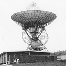 Black and white photo of antenna Deep Space Station 42 facing straight up, with a low building in front.