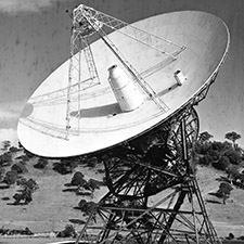 Black and white photo of antenna Deep Space Station 42 on an angle showing the dish, receiving cone and quad legs. Trees and hills in the background.