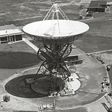 Aerial black and white photo of antenna Deep Space Station 42 facing straight up, with some buildings around it.