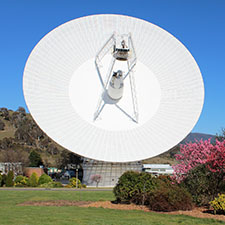 The antenna Deep Space Station 43 facing towards, showing the cone structure int eh middle and the quad legs. Grass and bushes in front with spring colours and blue sky behind.