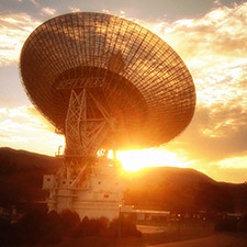 Sunset photo of Deep Space Station 43 antenna on a bit of an angle facing away. The image is red-yellow in colour from the setting Sun. Hills in the background, some clouds in the sky.