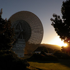 Sunset photo showing the back of Deep Space Station 43, and the Sun partially hidden by a tree. Grass and other trees a yellowy-colour from the setting Sun.