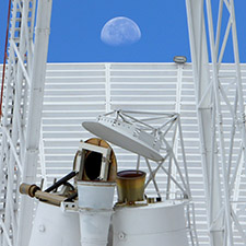 Feed horns of deep space station 43 in the foreground with part of the a leg on other side from the quad structure, the edge of the dish in the background with blue sky and a half Moon above it.