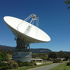 Deep SPace Station 43 on an angle showing the receiving cone and quad legs, a road in front, green grass, a green leafy tree and blue sky.