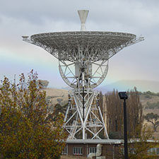 Antenna Deep Space Station 46 facing straight up with trees in the foreground, cloudy sky and a rainbow in the background.