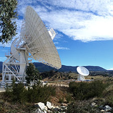 A rock bed, Deep Space Station 35 on an angle in the left of the photo and Deep Space Station 36 in the background. Hills behind and a blue sky with clouds.