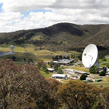 View of the Canberra Deep Space Communication Complex from a hill showing trees in the foreground, and the antennas and hills in the background. A mostly cloudy sky.