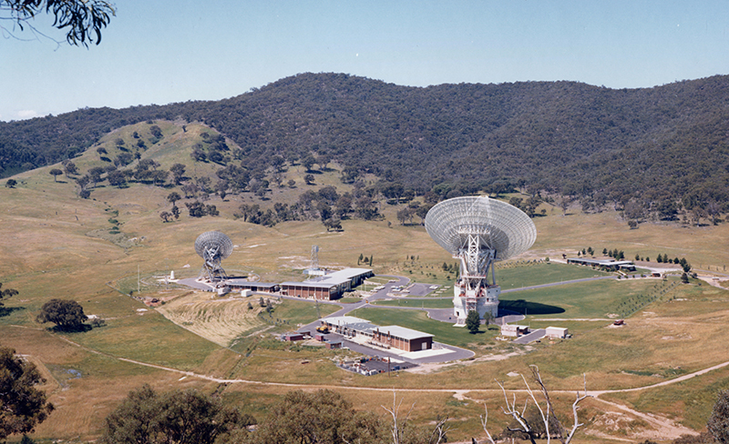 Hills covered in trees with a bare area in front with more buildings, some roads, and two antennas.