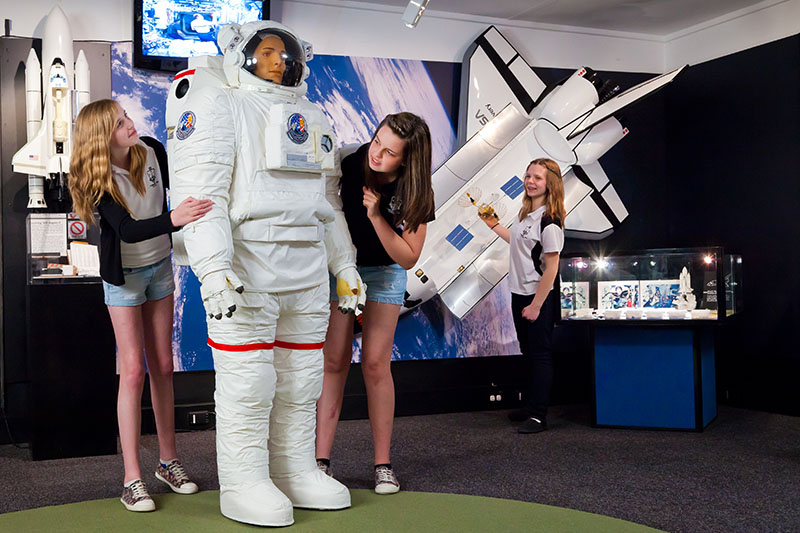 Two teenage girls looking at a model of a spacesuit while another girl stands next to a model of the space shuttle on the wall