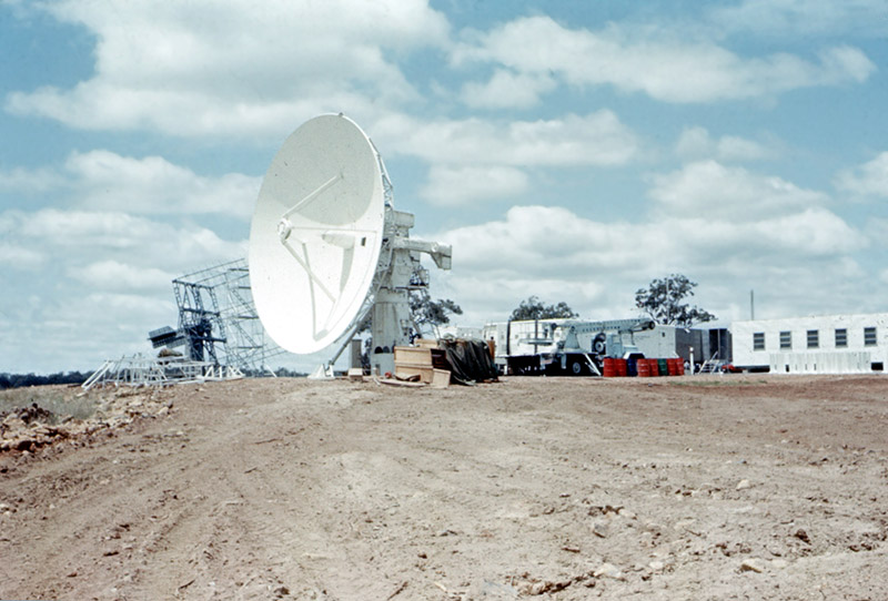 An antenna on an angle with dirt in front of it and trucks and building beside it, blue sky with clouds behind.