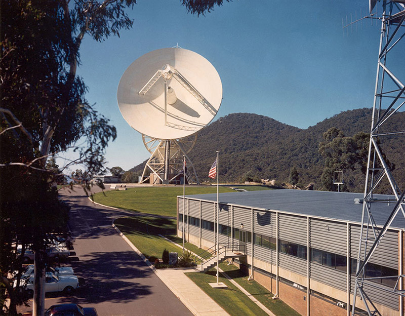 Side of a building with a road running beside it and an antenna in the background with the dish facing towards the camera and hills, Honeysuckle Creek