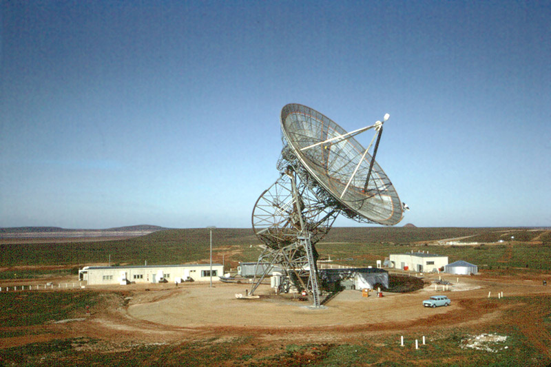 Antenna with the dish on an angle, a small number of buildings around it, and blue sky behind.