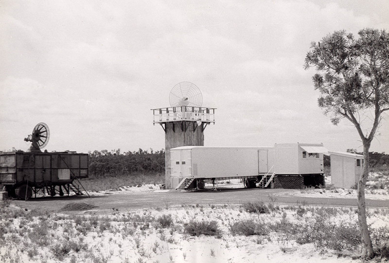 Black and white photo showing sand, trees, demountable type buildings and some small antenna dishes.